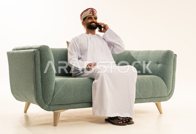 اجراء مكالمة هاتفية عبر الجوال، تطور التكنولوجيا والتقنيات الحديثة، ايماءات السعادة و السرور، بورتريه لرجل عربي خليجي عماني يرتدي الدشداشة و العمامة يجلس على الاريكة يقوم باستخدام الهاتف المحمول، خلفية بيضاء