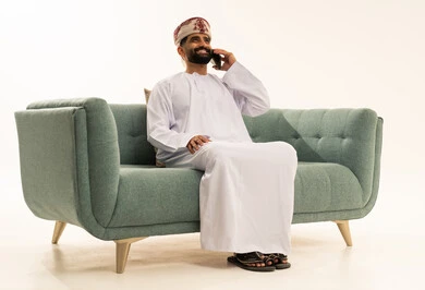 اجراء مكالمة هاتفية عبر الجوال، تطور التكنولوجيا والتقنيات الحديثة، ايماءات السعادة و السرور، بورتريه لرجل عربي خليجي عماني يرتدي الدشداشة و العمامة يجلس على الاريكة يقوم باستخدام الهاتف المحمول، خلفية بيضاء