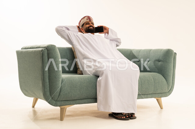 إيماءات الراحة والإسترخاء، اجراء مكالمة هاتفية عبر الجوال، تطور التكنولوجيا والتقنيات الحديثة، بورتريه لرجل عربي خليجي عماني يرتدي الدشداشة و العمامة يجلس على الاريكة يضع يديه خلف رأسه يقوم باستخدام الهاتف المحمول، خلفية بيضاء