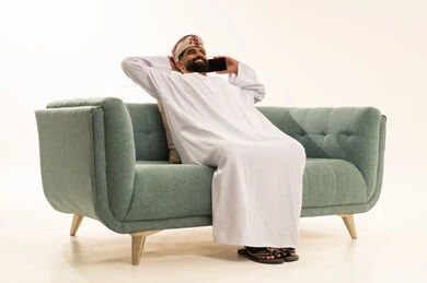 Gestures of comfort and relaxation, making a phone call via mobile, the evolution of technology and modern techniques, a portrait of an Arab Gulf Omani man wearing a dishdasha and a turban sitting on the sofa with his hands behind his head using a mobile phone, white background.