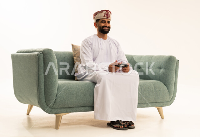Gestures of happiness and joy, interaction and immersion with PlayStation games, modern electronic entertainment technologies, spending enjoyable and entertaining time, a portrait of an Arab Gulf Omani man wearing a dishdasha and a turban sitting on the sofa holding a game controller playing video games, white background.
