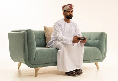 التفاعل والاندماج مع ألعاب البلايستيشن، تقنيات الكترونية حديثة ترفيهيه, قضاء وقت ممتع ومسلي، بورتريه لرجل عربي خليجي عماني يرتدي الدشداشة و العمامة يجلس على الاريكة يحمل بيده عصا التحكم يقوم باللعب في ألعاب الفيديو، خلفية بيضاء