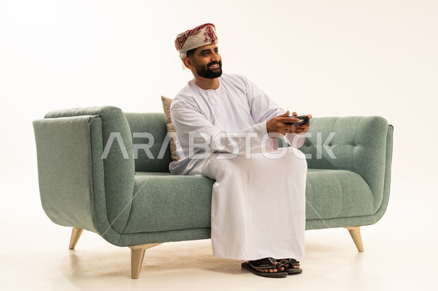 تقنيات الكترونية حديثة ترفيهيه, قضاء وقت ممتع ومسلي، التفاعل والاندماج مع ألعاب البلايستيشن، بورتريه لرجل عربي خليجي عماني يرتدي الدشداشة و العمامة يجلس على الاريكة يحمل بيده عصا التحكم يقوم باللعب في ألعاب الفيديو، خلفية بيضاء