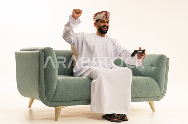 Gestures of victory and winning, interaction and immersion with PlayStation games, modern electronic entertainment technologies, a portrait of an Arab Gulf Omani man wearing a dishdasha and a turban sitting on the sofa holding a game controller playing video games, white background.