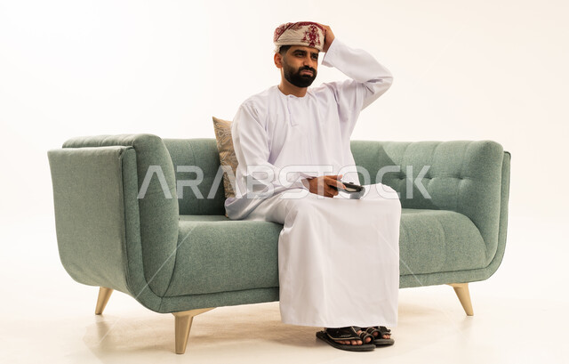 تعابير الاستمتاع باللعب بالألعاب الالكترونية الترفيهية، استخدام الأجهزة التكنولوجية والتقنيات الحديثة، بورتريه لرجل عربي خليجي عماني يرتدي الدشداشة و العمامة يجلس على الاريكة يمسك بيده عصا التحكم بإيماءات الخسارة ، خلفية بيضاء