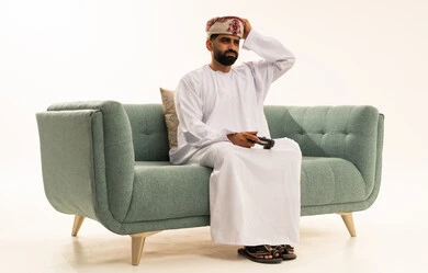 تعابير الاستمتاع باللعب بالألعاب الالكترونية الترفيهية، استخدام الأجهزة التكنولوجية والتقنيات الحديثة، بورتريه لرجل عربي خليجي عماني يرتدي الدشداشة و العمامة يجلس على الاريكة يمسك بيده عصا التحكم بإيماءات الخسارة ، خلفية بيضاء