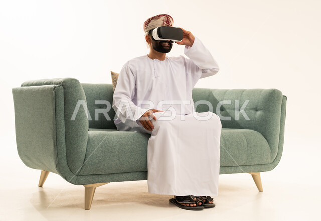 المتعة و التسلية بالمشاهدة عبر نظارة الواقع الافتراضي، التطور التقني والتكنولوجي، بورتريه لرجل عربي خليجي عماني يرتدي الدشداشة و العمامة يجلس على الاريكة  ينظر من خلال نظارات ثلاثية الأبعاد (الفي ار), الاندماج ومحاكاة العالم الخيالي، خلفية بيضاء