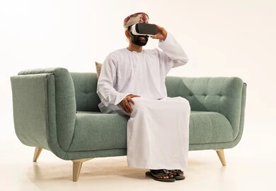 The enjoyment and entertainment of watching through virtual reality glasses, technological and technical development, a portrait of an Arab Gulf Omani man wearing a dishdasha and a turban sitting on a sofa looking through 3D glasses (VR), immersion and simulation of the imaginary world, white background.