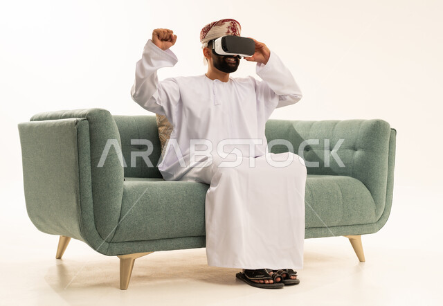رفع قبضة اليد بايماءات الانتصار، التطور التقني والتكنولوجي, بورتريه لرجل عربي خليجي عماني يرتدي الدشداشة و العمامة يجلس على الاريكة ينظر من خلال نظارات ثلاثية الأبعاد (الفي ار), الاندماج ومحاكاة العالم الخيالي، المتعة و التسلية بالمشاهدة عبر نظارة الواقع الافتراضي، خلفية بيضاء