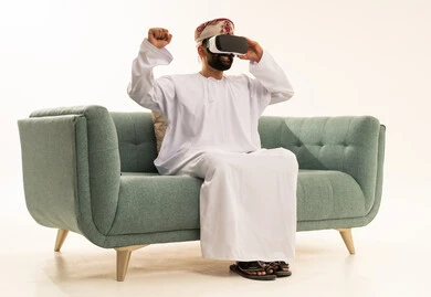 رفع قبضة اليد بايماءات الانتصار، التطور التقني والتكنولوجي, بورتريه لرجل عربي خليجي عماني يرتدي الدشداشة و العمامة يجلس على الاريكة ينظر من خلال نظارات ثلاثية الأبعاد (الفي ار), الاندماج ومحاكاة العالم الخيالي، المتعة و التسلية بالمشاهدة عبر نظارة الواقع الافتراضي، خلفية بيضاء