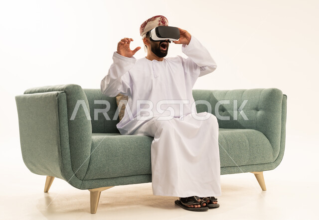 التطور التقني والتكنولوجي, بورتريه لرجل عربي خليجي عماني يرتدي الدشداشة و العمامة يجلس على الاريكة ينظر من خلال نظارات ثلاثية الأبعاد (الفي ار), الاندماج ومحاكاة العالم الخيالي، تعابير الاندهاش والتفاجؤ، المتعة و التسلية بالمشاهدة عبر نظارة الواقع الافتراضي، خلفية بيضاء