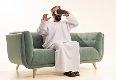 Technological and technical development, a portrait of an Arab Gulf Omani man wearing a dishdasha and turban sitting on a sofa looking through 3D glasses (VR), immersion and simulation of the imaginary world, expressions of amazement and surprise, enjoyment and entertainment while watching through virtual reality glasses, white background.