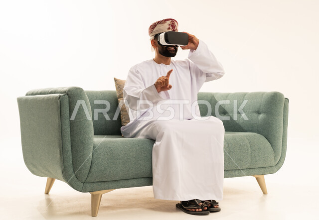 Seeing imaginary scenes, pointing with the index finger at something, using modern technological devices, interacting and merging with virtual reality, a portrait of an Arab Gulf Omani man wearing a dishdasha and turban sitting on a sofa wearing VR glasses, spending leisure time in an enjoyable way, white background.