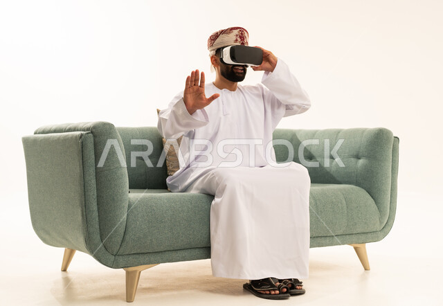 The enjoyment of watching through virtual reality glasses, technological and technical development, a portrait of an Arab Gulf Omani man wearing a dishdasha and a turban looking through 3D glasses (VR), raising his hand upwards with gestures of immersion and simulating the imaginary world, white background.