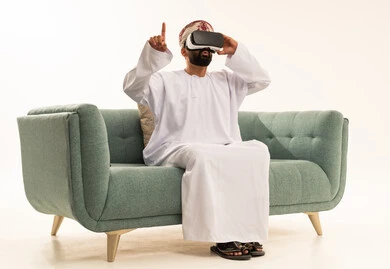الاشارة بإصبع السبابة إلى شيء ما، استخدام الأجهزة التقنية الحديثة، التفاعل والاندماج مع الواقع الافتراضي، رؤية المشاهد الخيالية، بورتريه لرجل عربي خليجي عماني يرتدي الدشداشة و العمامة يجلس على الاريكة يضع نظارات الفي آر، قضاء أوقات الفراغ بشكل ممتع، خلفية بيضاء