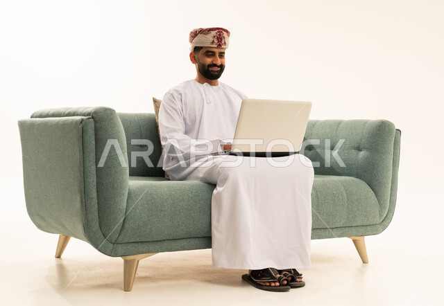 The concept of accomplishing remote work tasks, using modern and advanced technological devices, a portrait of an Arab Gulf Omani man wearing a dishdasha and a turban sitting on a sofa using a laptop, white background.
