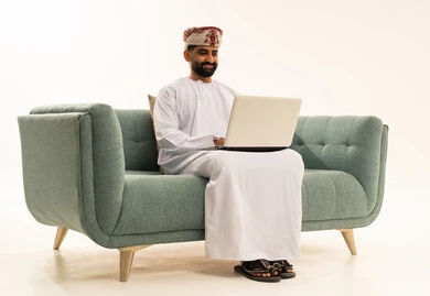 The concept of accomplishing remote work tasks, using modern and advanced technological devices, a portrait of an Arab Gulf Omani man wearing a dishdasha and a turban sitting on a sofa using a laptop, white background.