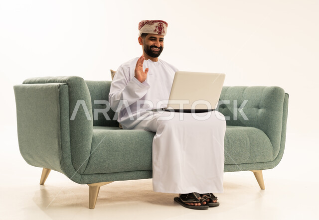 اجراء مكالمة فيديو عبر الانترنت، رفع كف اليد بتعابير التحية و السرور، مفهوم إنجاز مهام العمل عن بعد، استخدام الأجهزة التقنية الحديثة والمتطورة, بورتريه  لرجل عربي خليجي عماني يرتدي الدشداشة و العمامة يجلس على الاريكة يقوم باستعمال جهاز الحاسوب المحمول, خلفية بيضاء