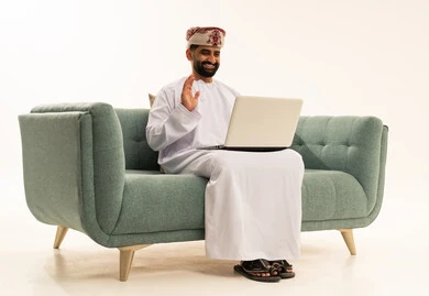 Making a video call over the internet, raising the hand with expressions of greeting and joy, the concept of completing work tasks remotely, using modern and advanced technological devices, a portrait of an Arab Gulf Omani man wearing a dishdasha and turban sitting on a sofa using a laptop, white background.