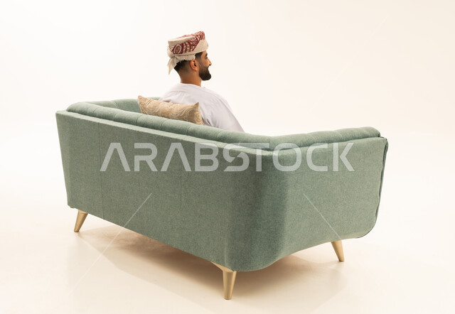 Enjoying and relaxing in a calm atmosphere, a portrait from behind of an Arab Gulf Omani man wearing a dishdasha and a turban sitting on a sofa looking at something, white background.