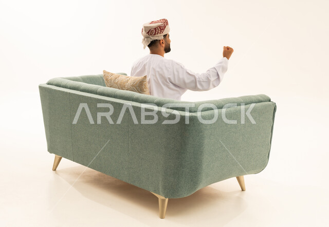 مفهوم الانجاز وتحقيق الاهداف، الانتصار والفوز والنجاح، بورتريه من الخلف لرجل عربي خليجي عماني يرتدي الدشداشة و العمامة يجلس على الاريكة يرفع قبضته للاعلى بإيماءات النصر، خلفية بيضاء