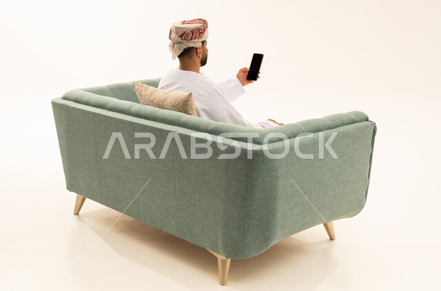 التواصل و الدردشة مع الاصدقاء، إجراء بث مباشر، بورتريه من الخلف لرجل عربي خليجي عماني يرتدي الدشداشة و العمامة يجلس على الاريكة يمسك بيده الهاتف المحمول، اجراء مكالمة فيديو ، خلفية بيضاء