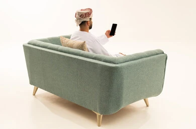 التواصل و الدردشة مع الاصدقاء، إجراء بث مباشر، بورتريه من الخلف لرجل عربي خليجي عماني يرتدي الدشداشة و العمامة يجلس على الاريكة يمسك بيده الهاتف المحمول، اجراء مكالمة فيديو ، خلفية بيضاء