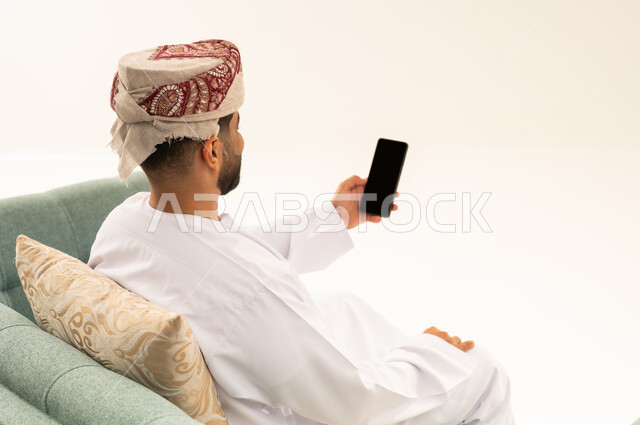 إجراء بث مباشر، التواصل و الدردشة مع الاصدقاء ، بورتريه من الخلف لرجل عربي خليجي عماني يرتدي الدشداشة و العمامة يجلس على الاريكة يمسك بيده الهاتف المحمول، اجراء مكالمة فيديو ، خلفية بيضاء