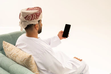 إجراء بث مباشر، التواصل و الدردشة مع الاصدقاء ، بورتريه من الخلف لرجل عربي خليجي عماني يرتدي الدشداشة و العمامة يجلس على الاريكة يمسك بيده الهاتف المحمول، اجراء مكالمة فيديو ، خلفية بيضاء