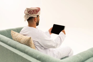 استعمال التكنولوجيا الحديثة والمتطورة للتعلم، تصفح تطبيقات جهاز التابلت، بورتريه من الخلف لرجل عربي خليجي عماني يرتدي الدشداشة و العمامة يجلس على الاريكة يقوم بمتابعة اخبار وسائل التواصل الاجتماعي باستخدام التابلت، خلفية بيضاء