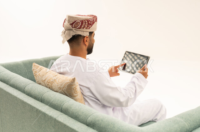محادثات ودردشات عبر الانترنت، تصفح تطبيقات جهاز التابلت، بورتريه من الخلف لرجل عربي خليجي عماني يرتدي الدشداشة و العمامة يجلس على الاريكة يقوم بمتابعة اخبار وسائل التواصل الاجتماعي باستخدام التابلت، خلفية بيضاء