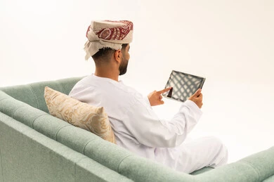 محادثات ودردشات عبر الانترنت، تصفح تطبيقات جهاز التابلت، بورتريه من الخلف لرجل عربي خليجي عماني يرتدي الدشداشة و العمامة يجلس على الاريكة يقوم بمتابعة اخبار وسائل التواصل الاجتماعي باستخدام التابلت، خلفية بيضاء