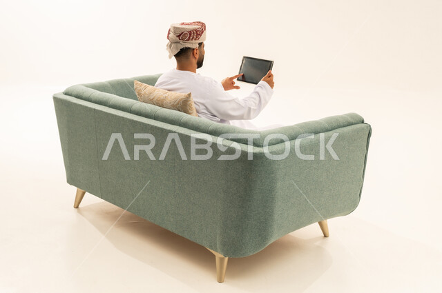الدردشة والتحدث مع الاصدقاء عبر الانترنت، تصفح تطبيقات جهاز التابلت، بورتريه من الخلف لرجل عربي خليجي عماني يرتدي الدشداشة و العمامة يجلس على الاريكة يقوم بمتابعة اخبار وسائل التواصل الاجتماعي باستخدام التابلت، خلفية بيضاء