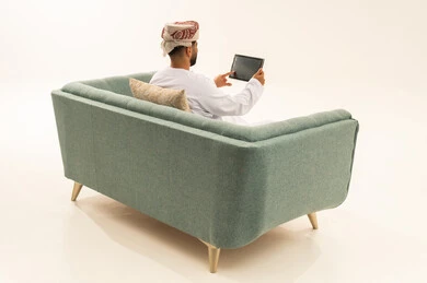 الدردشة والتحدث مع الاصدقاء عبر الانترنت، تصفح تطبيقات جهاز التابلت، بورتريه من الخلف لرجل عربي خليجي عماني يرتدي الدشداشة و العمامة يجلس على الاريكة يقوم بمتابعة اخبار وسائل التواصل الاجتماعي باستخدام التابلت، خلفية بيضاء