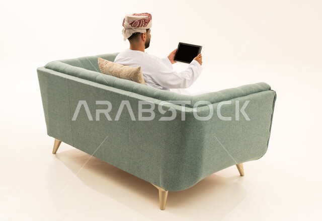 تصفح تطبيقات جهاز التابلت، الدردشة والتحدث مع الاصدقاء عبر الانترنت، بورتريه من الخلف لرجل عربي خليجي عماني يرتدي الدشداشة و العمامة يجلس على الاريكة يقوم بمتابعة اخبار وسائل التواصل الاجتماعي باستخدام التابلت، خلفية بيضاء