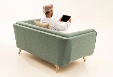 تصفح تطبيقات جهاز التابلت، الدردشة والتحدث مع الاصدقاء عبر الانترنت، بورتريه من الخلف لرجل عربي خليجي عماني يرتدي الدشداشة و العمامة يجلس على الاريكة يقوم بمتابعة اخبار وسائل التواصل الاجتماعي باستخدام التابلت، خلفية بيضاء