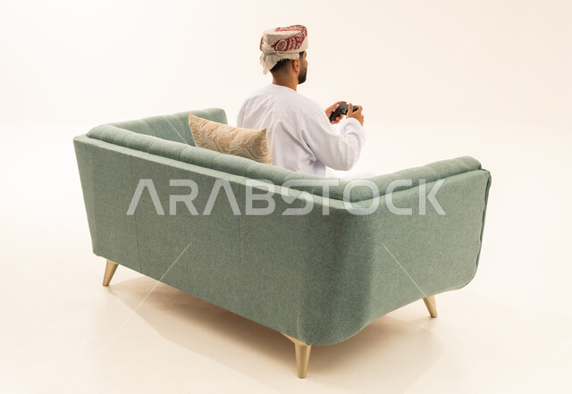 قضاء أوقات ممتعة في ألعاب الفيديو الترفيهيه، بورتريه من الخلف لرجل عربي خليجي عماني يرتدي الدشداشة و العمامة يجلس على الاريكة يحمل بيده عصا التحكم، إيماءات الاستمتاع والاندماج، استخدام الأجهزة التكنولوجية والتقنيات الحديثة، خلفية بيضاء