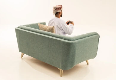 Spending enjoyable times in recreational video games, a portrait from behind of an Arab Gulf Omani man wearing a dishdasha and a turban sitting on the sofa holding a game controller, gestures of enjoyment and immersion, using technological devices and modern techniques, white background.