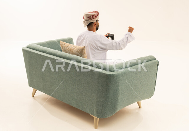 رفع قبضة اليد للأعلى بتعابير الانتصار والفوز، إيماءات الاستمتاع والاندماج والتفاعل, استخدام الأجهزة التكنولوجية والتقنيات الحديثة، بورتريه من الخلف لرجل عربي خليجي عماني يرتدي الدشداشة و العمامة يجلس على الاريكة يمسك بيده عصا التحكم، اللعب بألعاب الفيديو الترفيهية، خلفية بيضاء