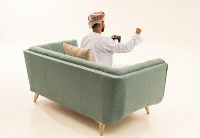 Raising a fist upwards with expressions of victory and triumph, gestures of enjoyment, engagement, and interaction, using technological devices and modern techniques, a portrait from behind of an Arab Gulf Omani man wearing a dishdasha and turban sitting on a sofa holding a game controller, playing recreational video games, white background.