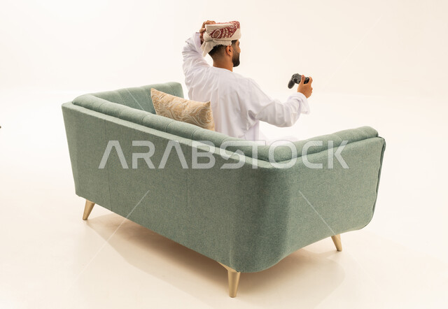 إيماءات الخسارة وخيبة الأمل، استخدام الألعاب الإلكترونية والأجهزة الحديثة، بورتريه من الخلف لرجل عربي خليجي عماني يرتدي الدشداشة و العمامة يجلس على الاريكة يمسك عصا التحكم يضع يده على رأسه بتعابير الحسرة والحزن، التسلية والترفيه بالفيديو جيمز في وقت الفراغ، خلفية بيضاء