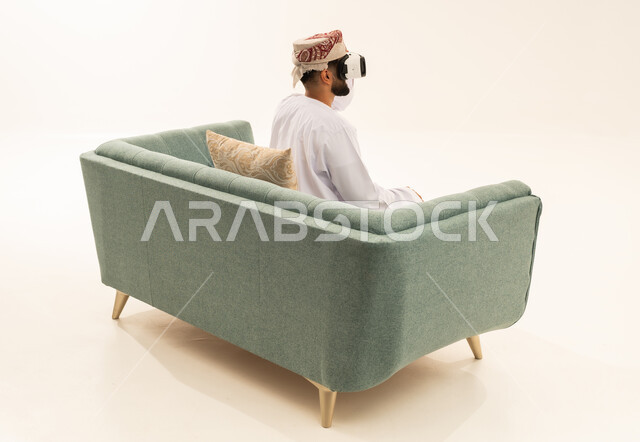 Integrating modern technologies and technology with fun and entertainment, simulating the imaginary world, a portrait from behind of an Arab Gulf Omani man wearing a dishdasha and a turban sitting on a sofa wearing 3D virtual reality glasses, gestures of immersion and focus, white background.