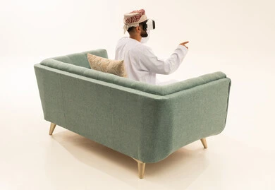 التطور التقني والتكنولوجي ، الاشارة الى شيء ما، المتعة والتسلية بالمشاهدة عبر نظارة الواقع الافتراضي، الاندماج ومحاكاة العالم الخيالي، بورتريه من الخلف لرجل عربي خليجي عماني يرتدي الدشداشة و العمامة يجلس على الاريكة يضع نظارات ثلاثية الأبعاد (الفي ار)، خلفية بيضاء