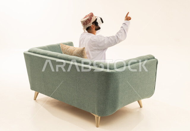 Referring to something, the enjoyment and entertainment of watching through virtual reality glasses, immersion and simulation of the imaginary world, a portrait from behind of an Arab Gulf Omani man wearing a dishdasha and turban sitting on a sofa wearing 3D glasses (VR), technological and technical development, white background.