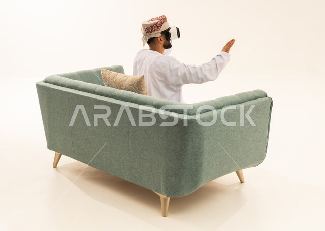 المتعة بالمشاهدة عبر نظارة الواقع الافتراضي، التطور التقني والتكنولوجي, بورتريه من الخلف لرجل عربي خليجي عماني يرتدي الدشداشة و العمامة يجلس على الاريكة ينظر من خلال نظارات ثلاثية الأبعاد (الفي ار), رفع كف اليد للأعلى بإيماءات الاندماج ومحاكاة العالم الخيالي، خلفية بيضاء