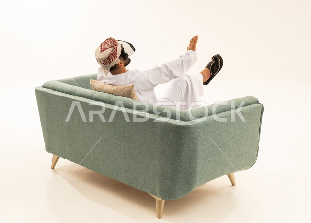 Entertainment through virtual reality glasses technology, gestures of immersion and interaction with the world of imagination, a portrait from behind of an Arab Gulf Omani man wearing a dishdasha and a turban sitting on a sofa wearing 3D glasses (VR), technological and technical development, white background.
