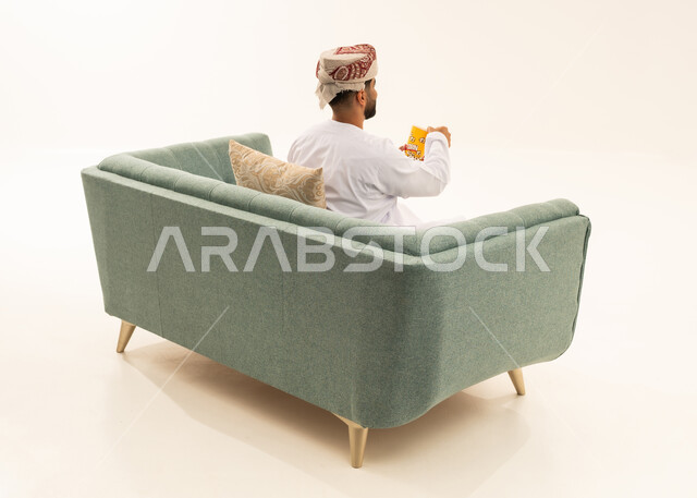 الترفيه عن النفس وقضاء وقت ممتع، الاستمتاع باكل البوب كورن، سناكس ومقرمشات لذيذة، انشطة ترفيهية منزلية، الاستمتاع بتناول الفشار، بورتريه من الخلف لرجل عربي خليجي عماني يرتدي الدشداشة و العمامة يجلس على الاريكة يحمل بيده علبة الذرة، تناول سناكات خفيفة وصحية،خلفيه بيضاء