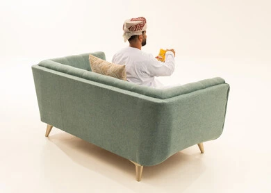 الترفيه عن النفس وقضاء وقت ممتع، الاستمتاع باكل البوب كورن، سناكس ومقرمشات لذيذة، انشطة ترفيهية منزلية، الاستمتاع بتناول الفشار، بورتريه من الخلف لرجل عربي خليجي عماني يرتدي الدشداشة و العمامة يجلس على الاريكة يحمل بيده علبة الذرة، تناول سناكات خفيفة وصحية،خلفيه بيضاء