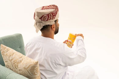 الاستمتاع باكل البوب كورن، سناكس ومقرمشات لذيذة، انشطة ترفيهية منزلية، الاستمتاع بتناول الفشار، بورتريه من الخلف لرجل عربي خليجي عماني يرتدي الدشداشة و العمامة يجلس على الاريكة يحمل بيده علبة الذرة، تناول سناكات خفيفة وصحية، الترفيه عن النفس وقضاء وقت ممتع،خلفيه بيضاء