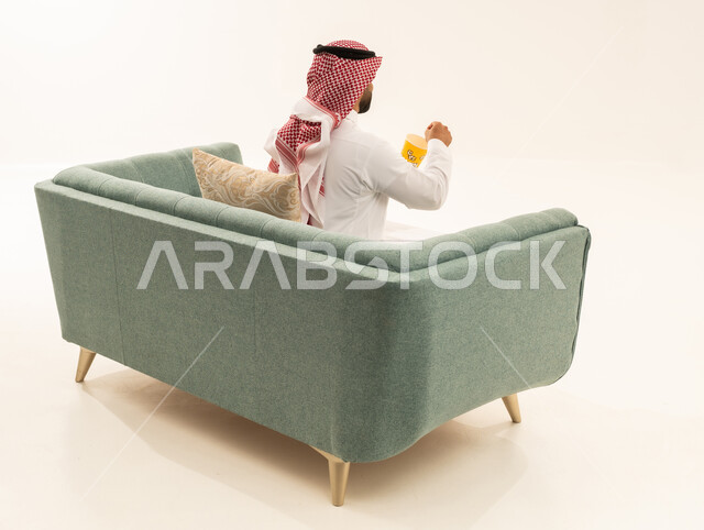 الاستمتاع بتناول الفشار، انشطة ترفيهية منزلية، بورتريه من الخلف لرجل عربي خليجي سعودي يرتدي الثوب التقليدي و الشماغ يجلس على الاريكة يحمل بيده علبة الذرة، سناكات ومقرمشات لذيذة، تناول سناكات خفيفة وصحية، الاستمتاع باكل البوب كورن، الترفيه عن النفس وقضاء وقت ممتع،خلفيه بيضاء